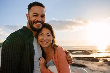 Man holding his partner with his arm around her standing near a beach at sunset. Man and woman enjoying vacation near the seashore.